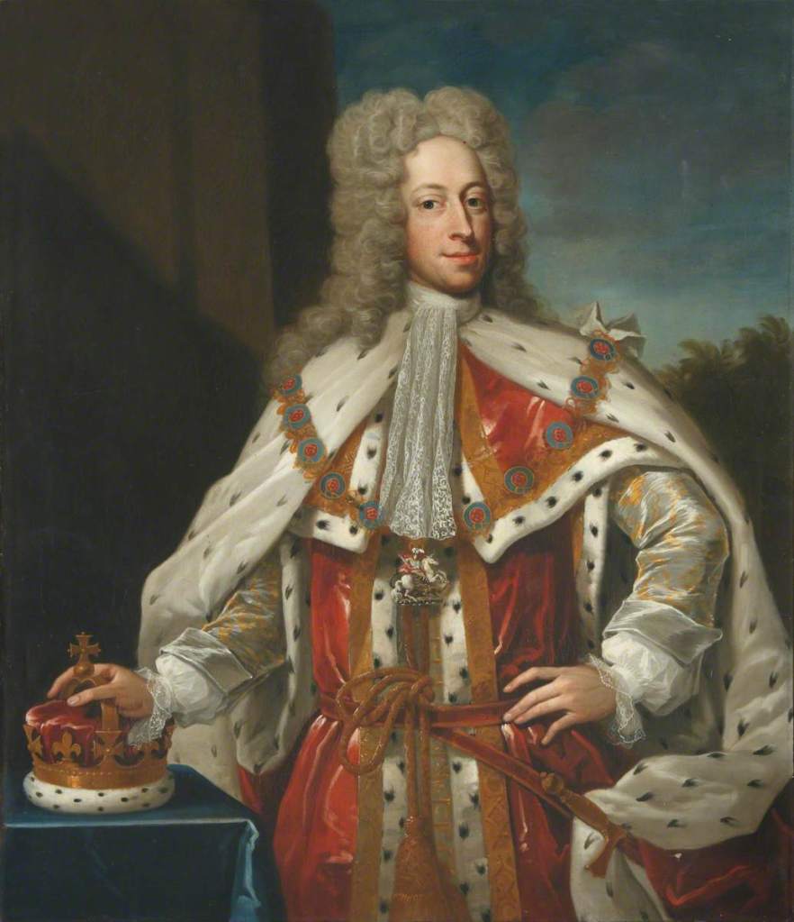 Painting of Prince Frederick. He is stood with his left hand on his hip and the other on a royal crown, which is resting on a table covered in a blue cloth. He is wearing a long curly grey wig, ermine robes of red and white, with a large gold necklace over his shoulders, and a white lace collar tied at his neck. 